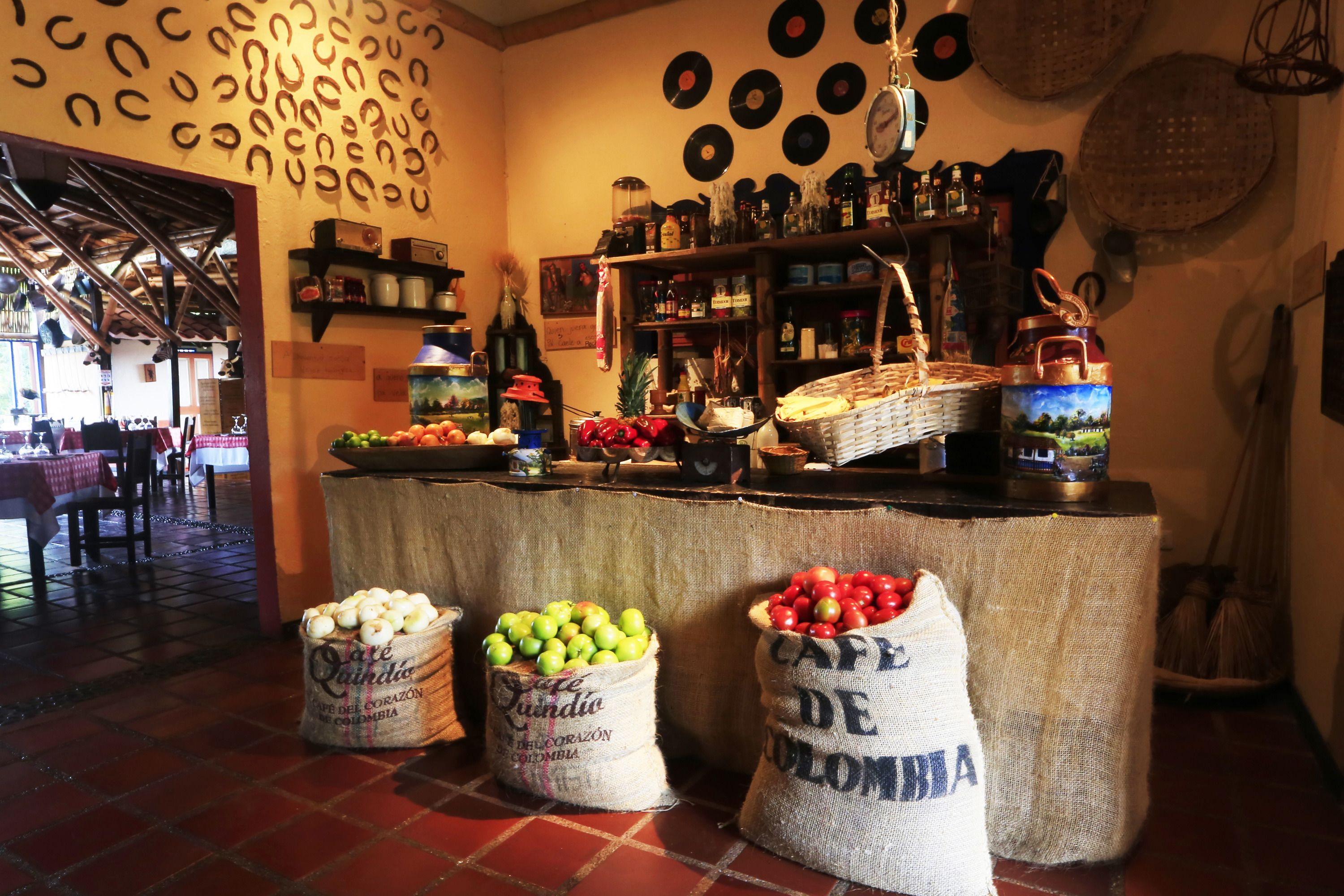 A bar area with a rustic decor, featuring a wooden ceiling, shelves with bottles, and a wall adorned with records. There are sacks of apples and a basket of bread on the counter.
