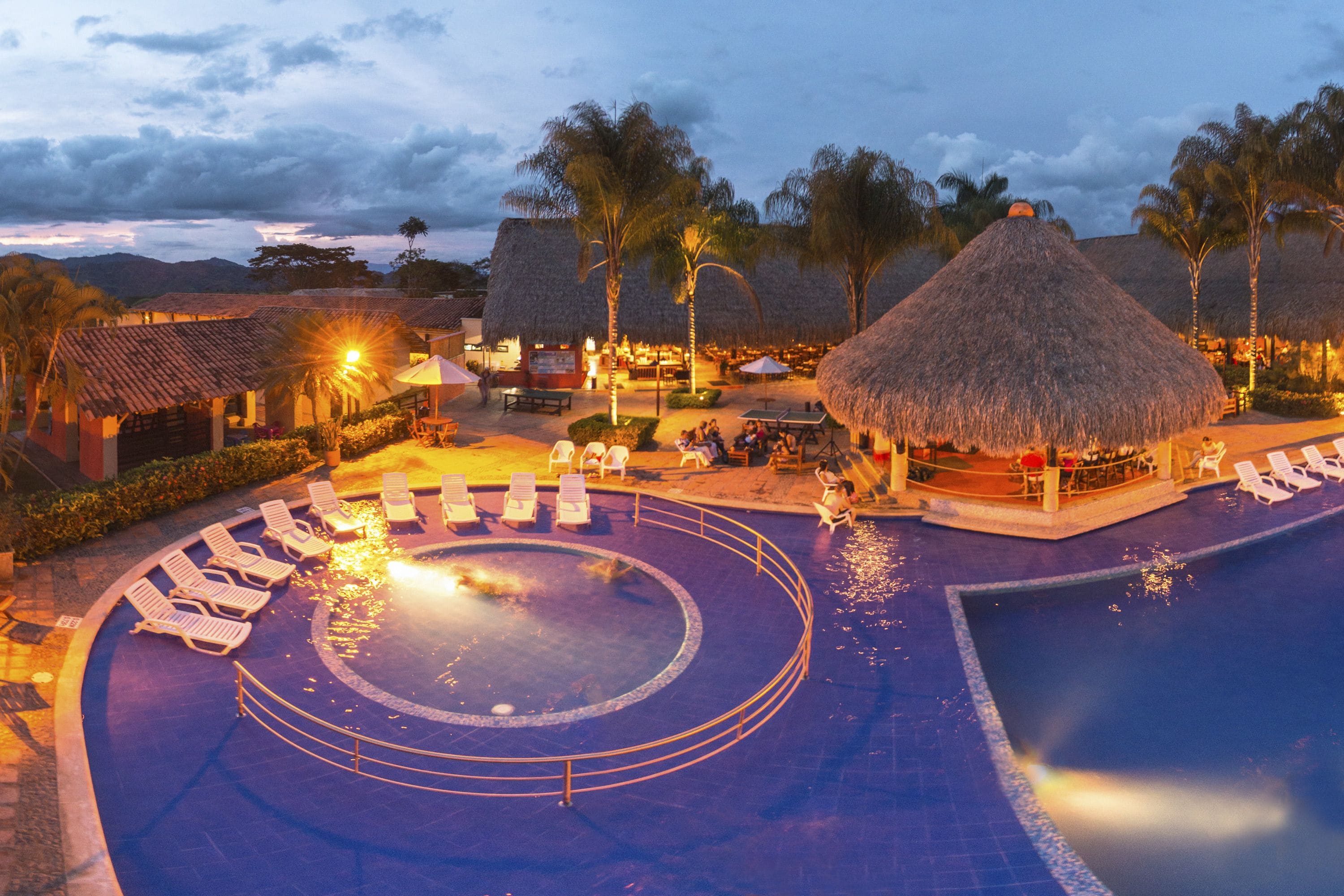 Area piscina di un resort con lettini, una vasca idromassaggio e strutture coperte da tetti di paglia.