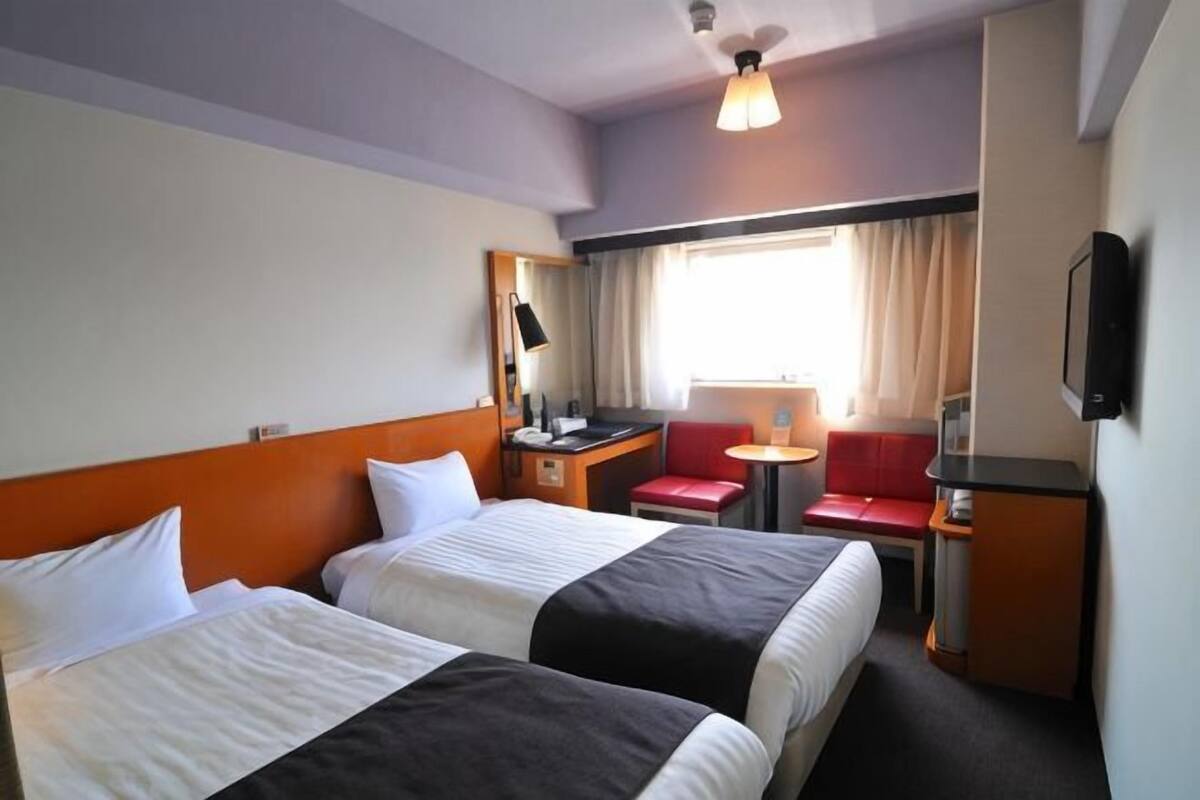 Une chambre d’hôtel avec deux lits, un bureau avec une chaise, une petite table, un téléviseur et une fenêtre avec des rideaux.