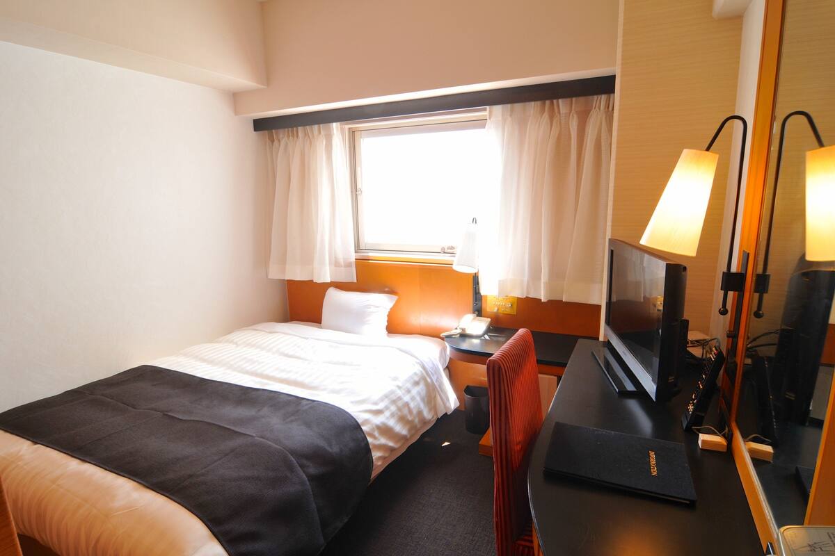 A hotel room with a bed, a desk with a computer, a TV, and a window with curtains.