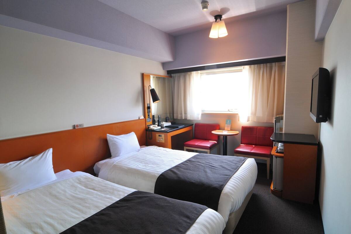A hotel room with two beds, a desk with a chair, a TV, and a window with curtains.