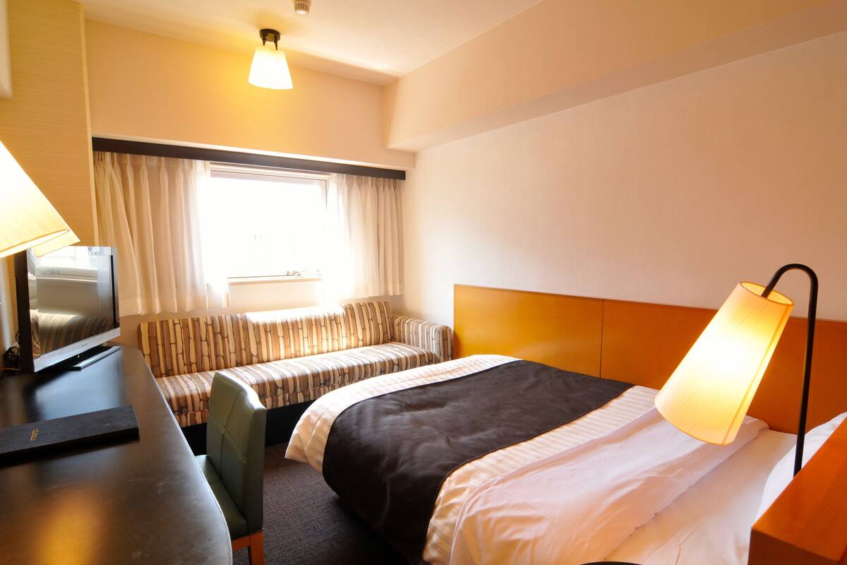 A hotel room with a bed, a sofa, a desk with a television, and a lamp.