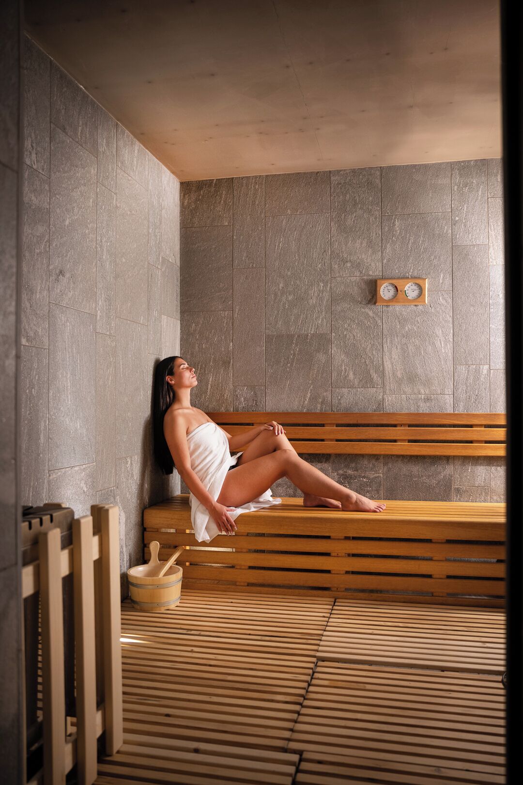 A person relaxing on a wooden bench in a sauna room with stone walls and a wooden floor.