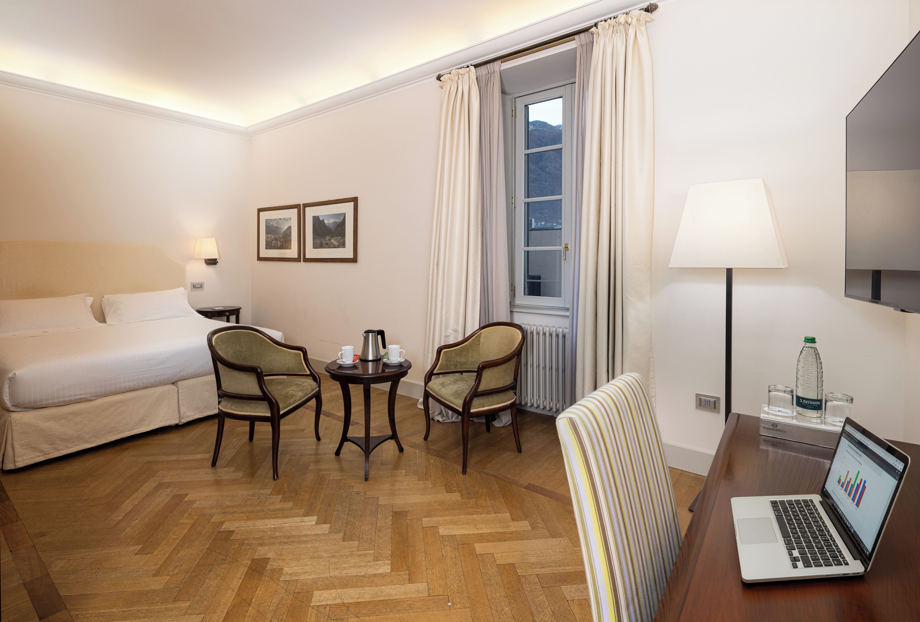 A hotel room with a bed, two armchairs, a small table with a coffee pot, a laptop, and a window with curtains.