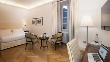 A hotel room with a bed, two armchairs, a small table with a coffee pot, a laptop, and a window with curtains.