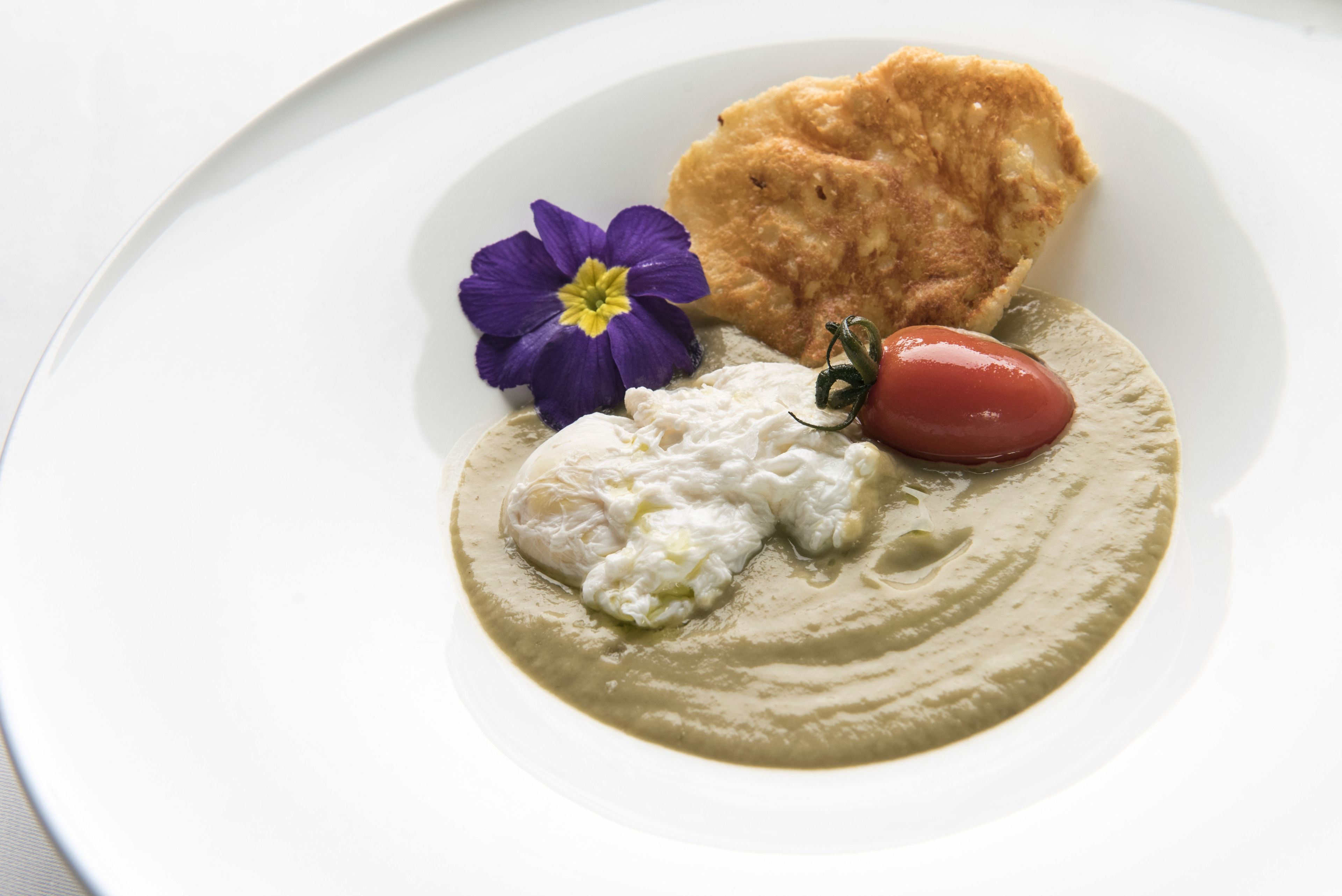 Un plato blanco con una cucharada de crema, un trozo de pan, una flor morada y un tomate pequeño rojo.