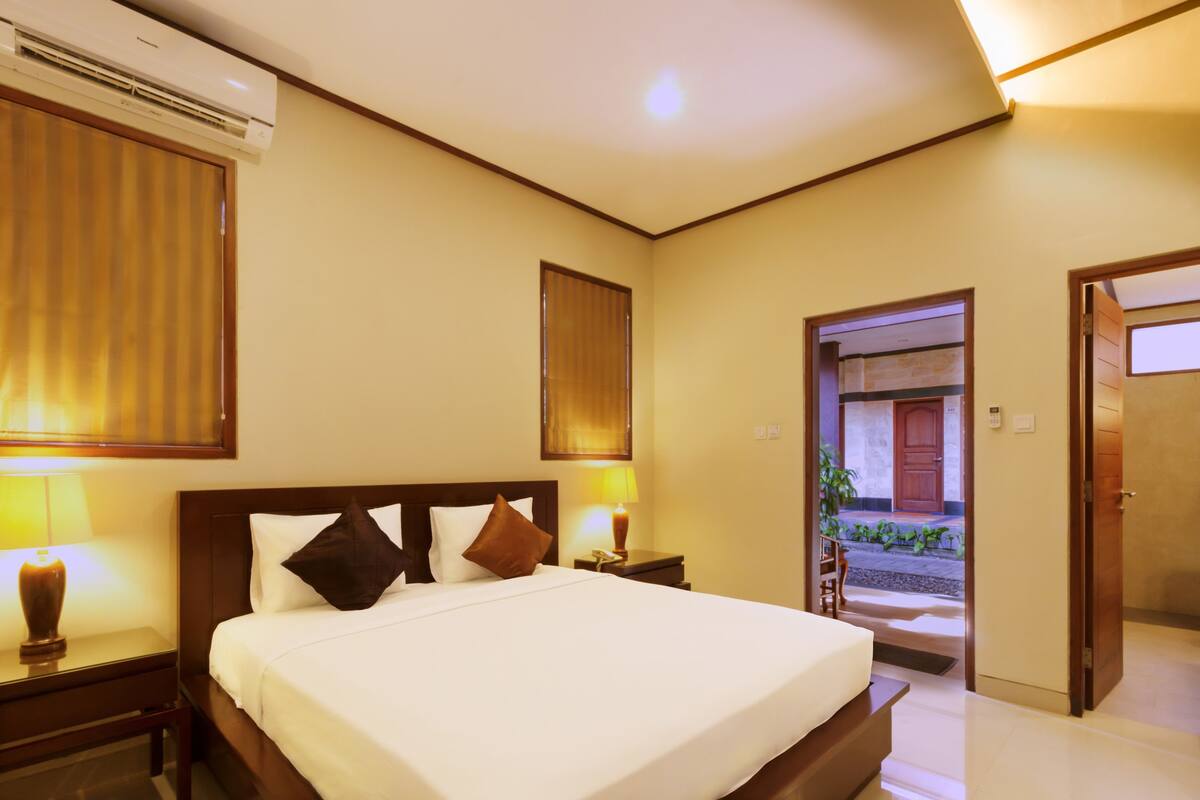 A hotel room with a large bed, bedside lamps, an air conditioning unit, and a view of the outside through a door.