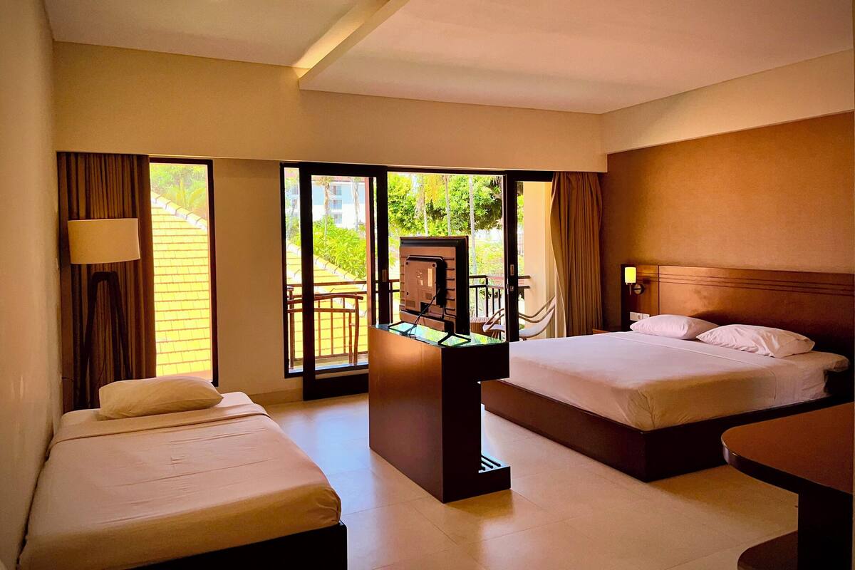 A hotel room with two beds, a desk, and a balcony with a view of greenery.