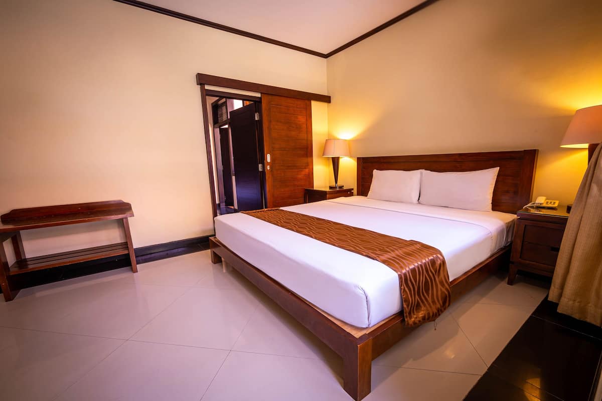 A hotel room with a large bed, a wooden bench, two bedside tables with lamps, and a door leading to another room.
