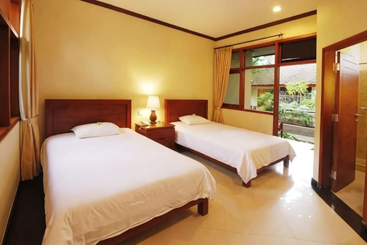 Habitación de hotel con dos camas, una mesita de noche con lámpara, cabecera de madera y vistas al exterior.