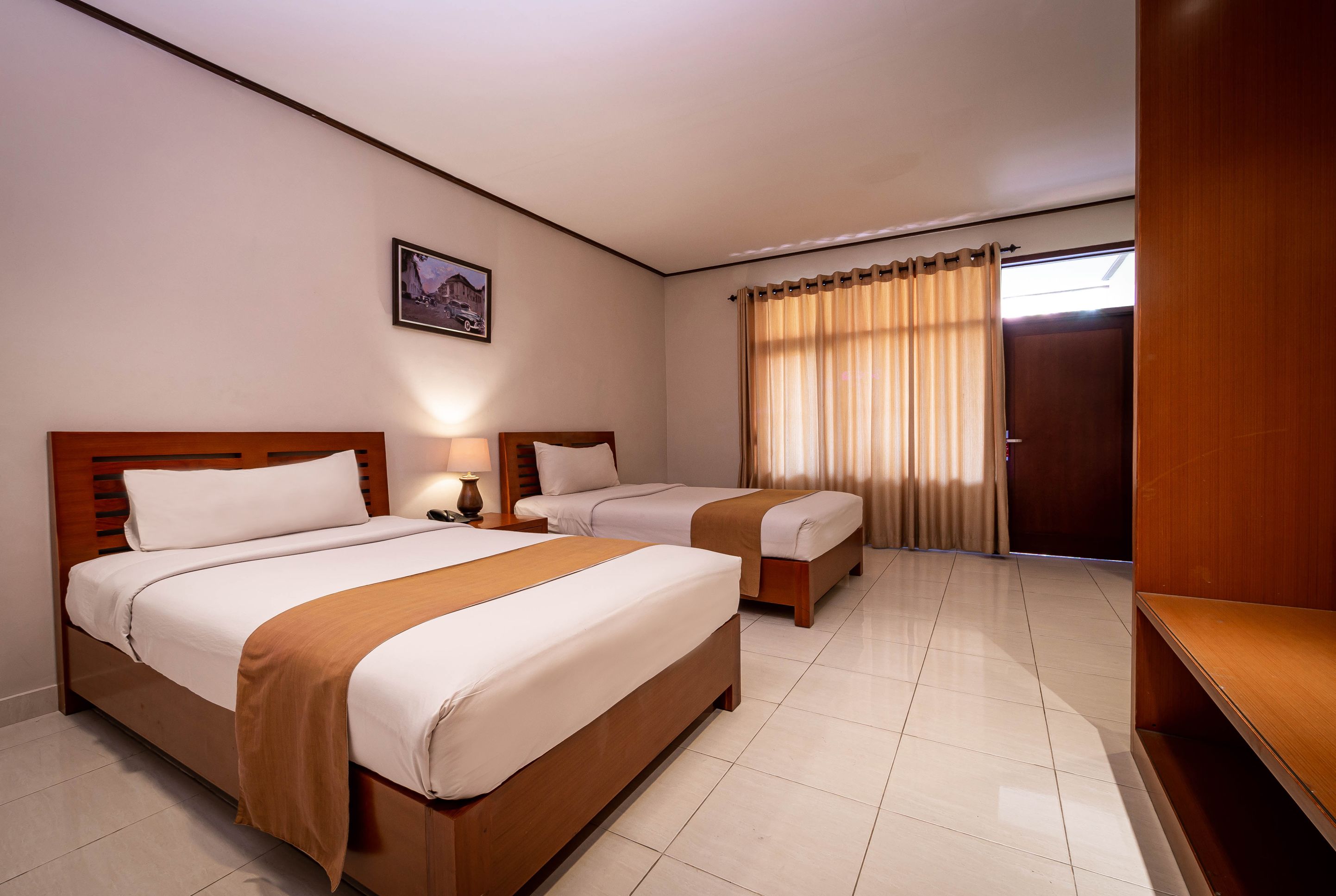 Quarto de hotel com duas camas, cabeceira de madeira, mesa de cabeceira com abajur, janela com cortinas e porta.