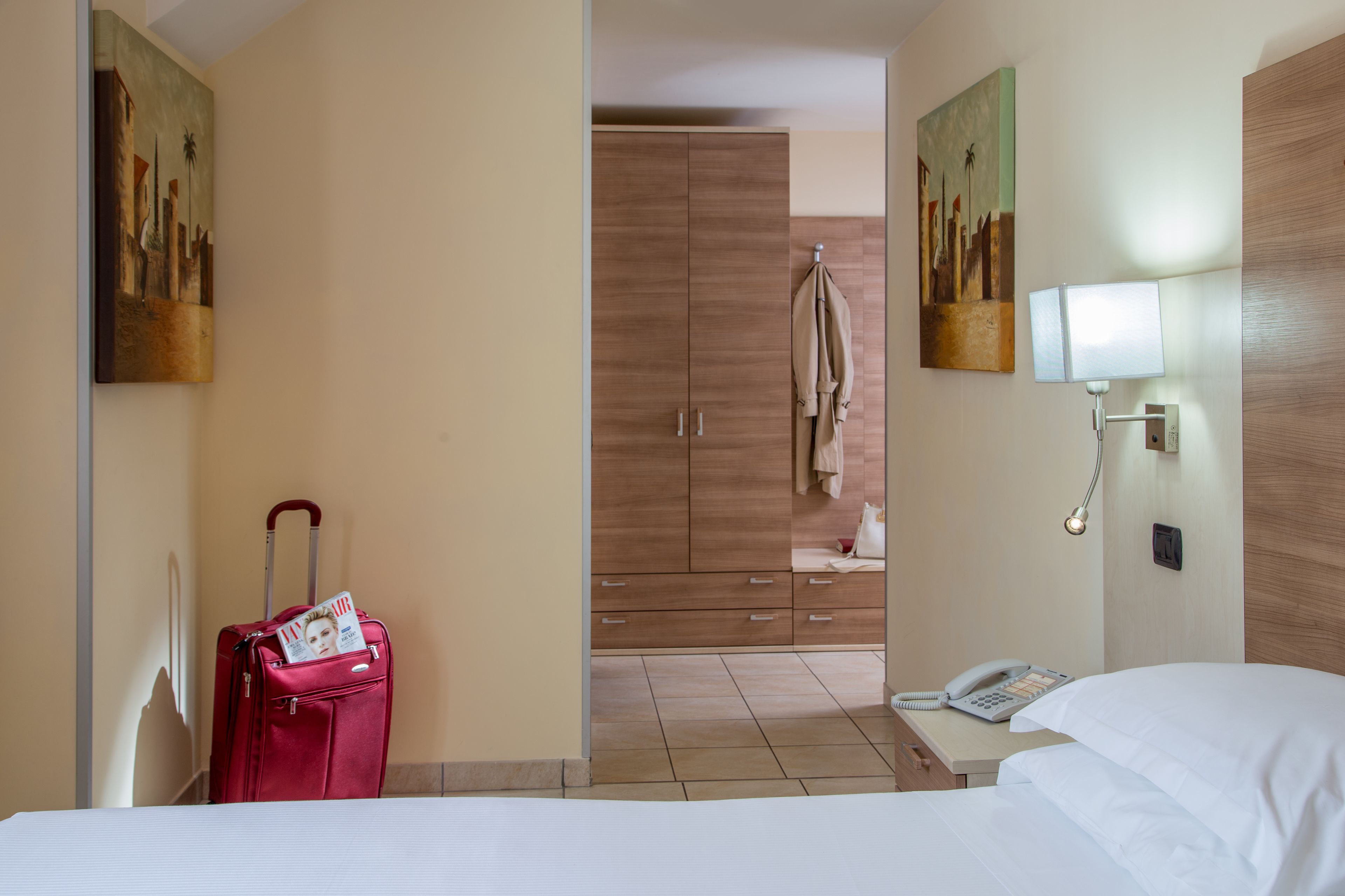 A hotel room with a bed, a desk with a telephone, a red suitcase, a wooden wardrobe, and two paintings on the walls.