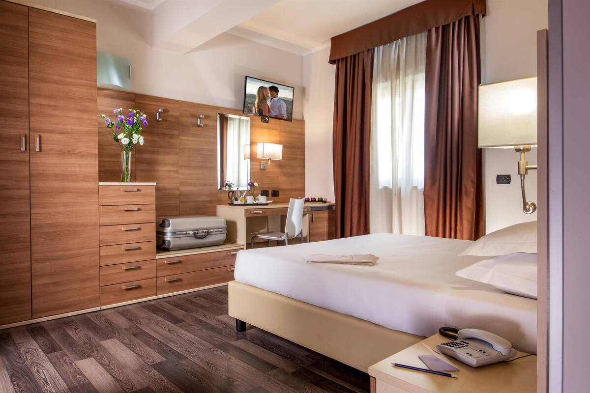 A hotel room with a large bed, wooden wardrobe, desk with a chair, a mirror, and a television on the wall.