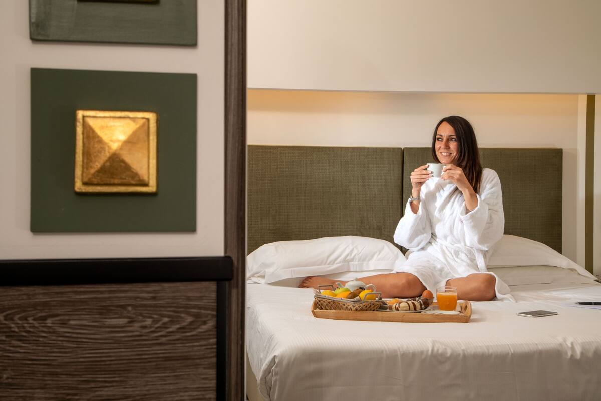 A person sitting on a bed with a tray of breakfast items, including fruits and juice, in a room with a green wall and a framed picture.