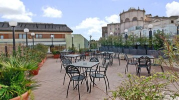 An outdoor patio area with tables and chairs, surrounded by potted plants and buildings in the background.
