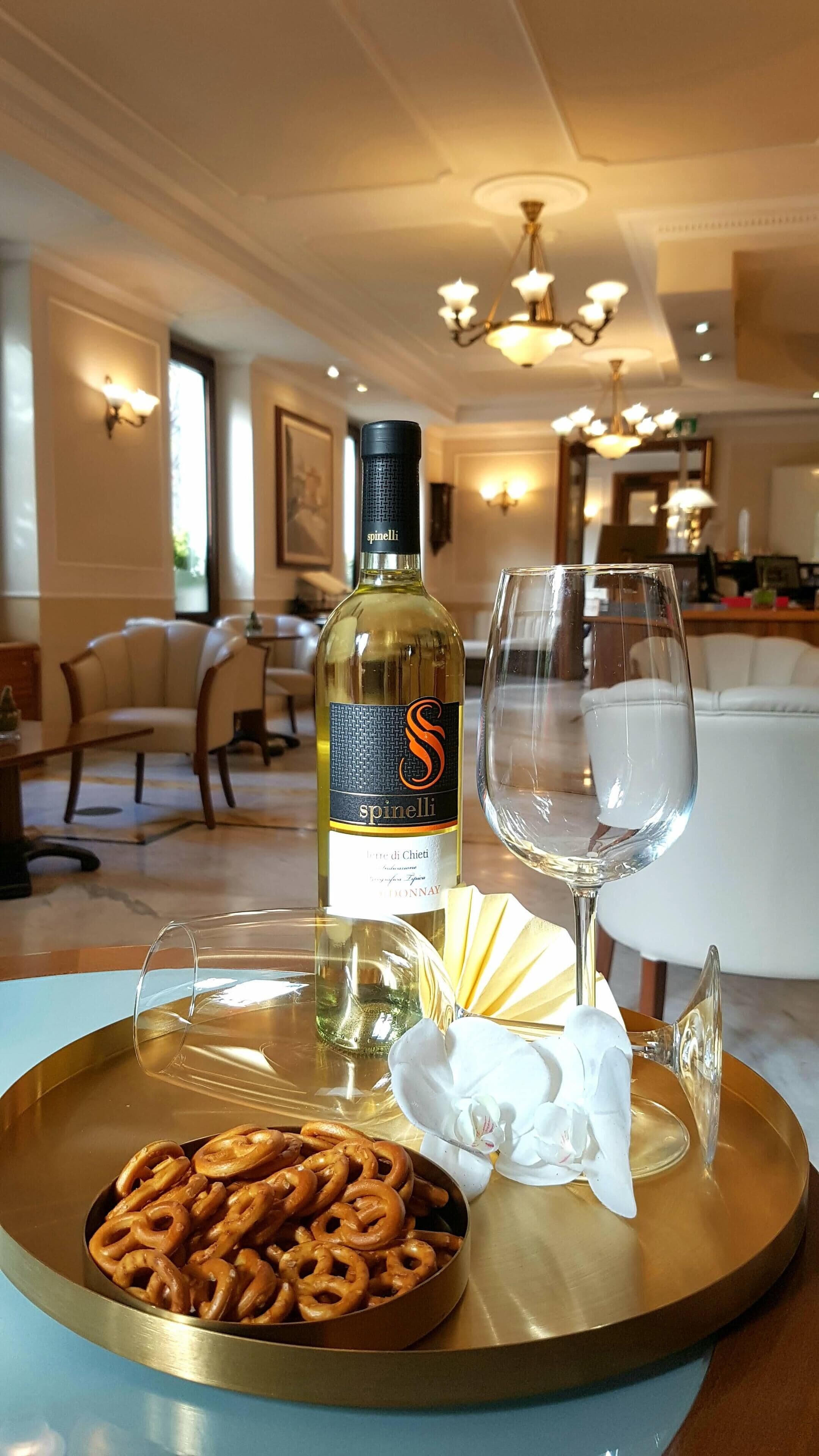 A bottle of wine, a glass, pretzels, and a fan on a tray in a dining area with a chandelier and a sofa in the background.