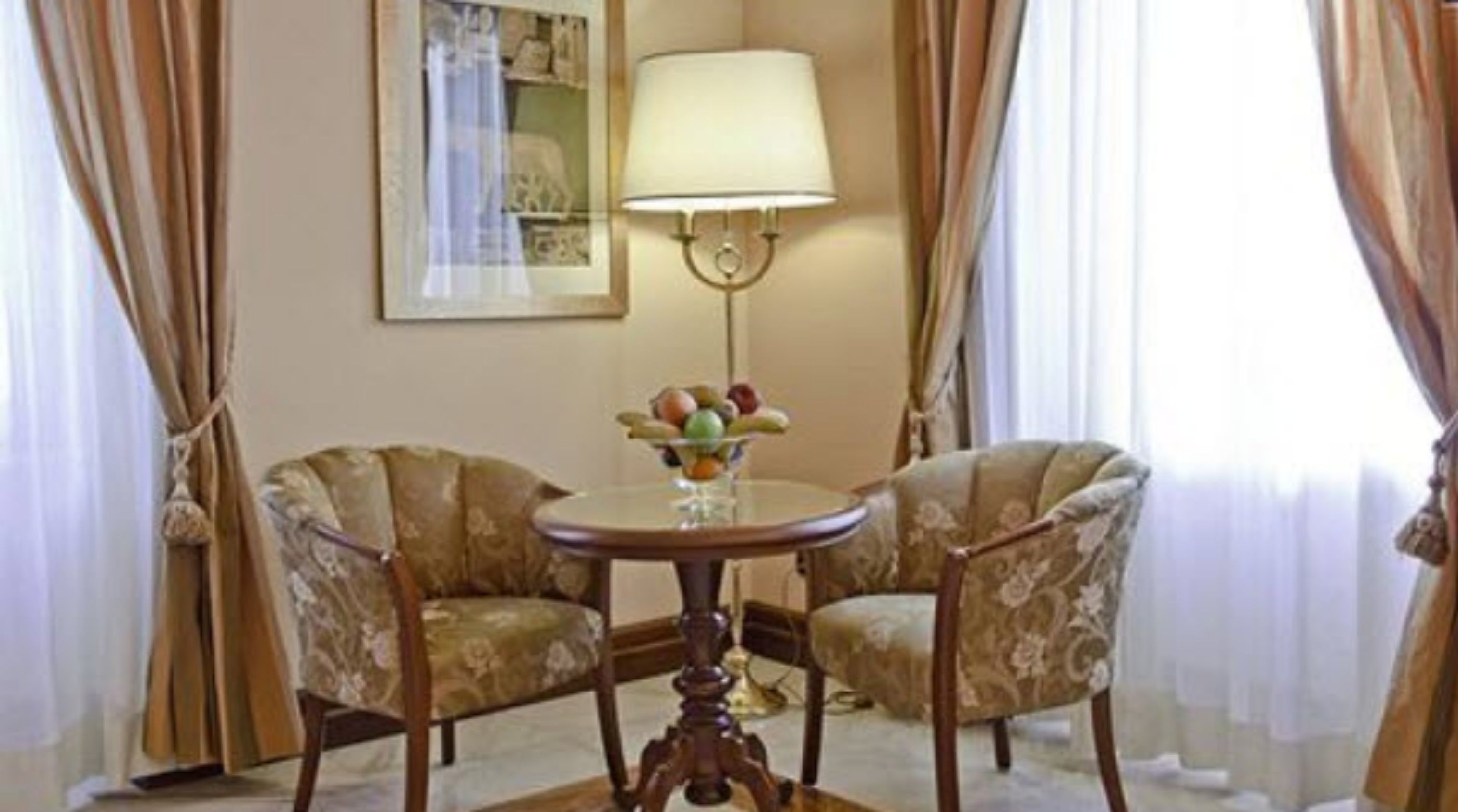 Ein Zimmer mit zwei Polstersesseln, einem runden Tisch mit einer Obstschale, einer Wandlampe und einem gerahmten Bild.