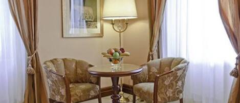 Una habitación con dos sillones tapizados, una mesa redonda con fruteros, una lámpara de pared e imagen enmarcadas.