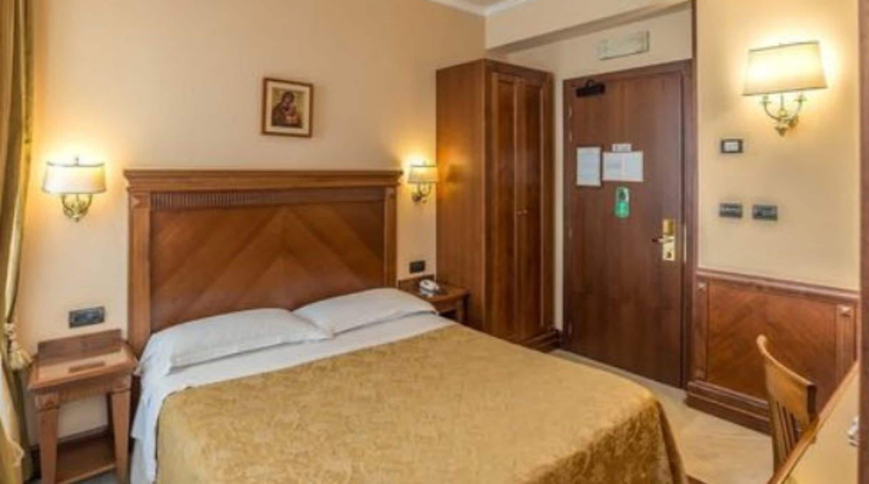 A hotel room with a large bed, wooden headboard, two bedside tables, a nightstand, a wardrobe, and two wall-mounted lamps.