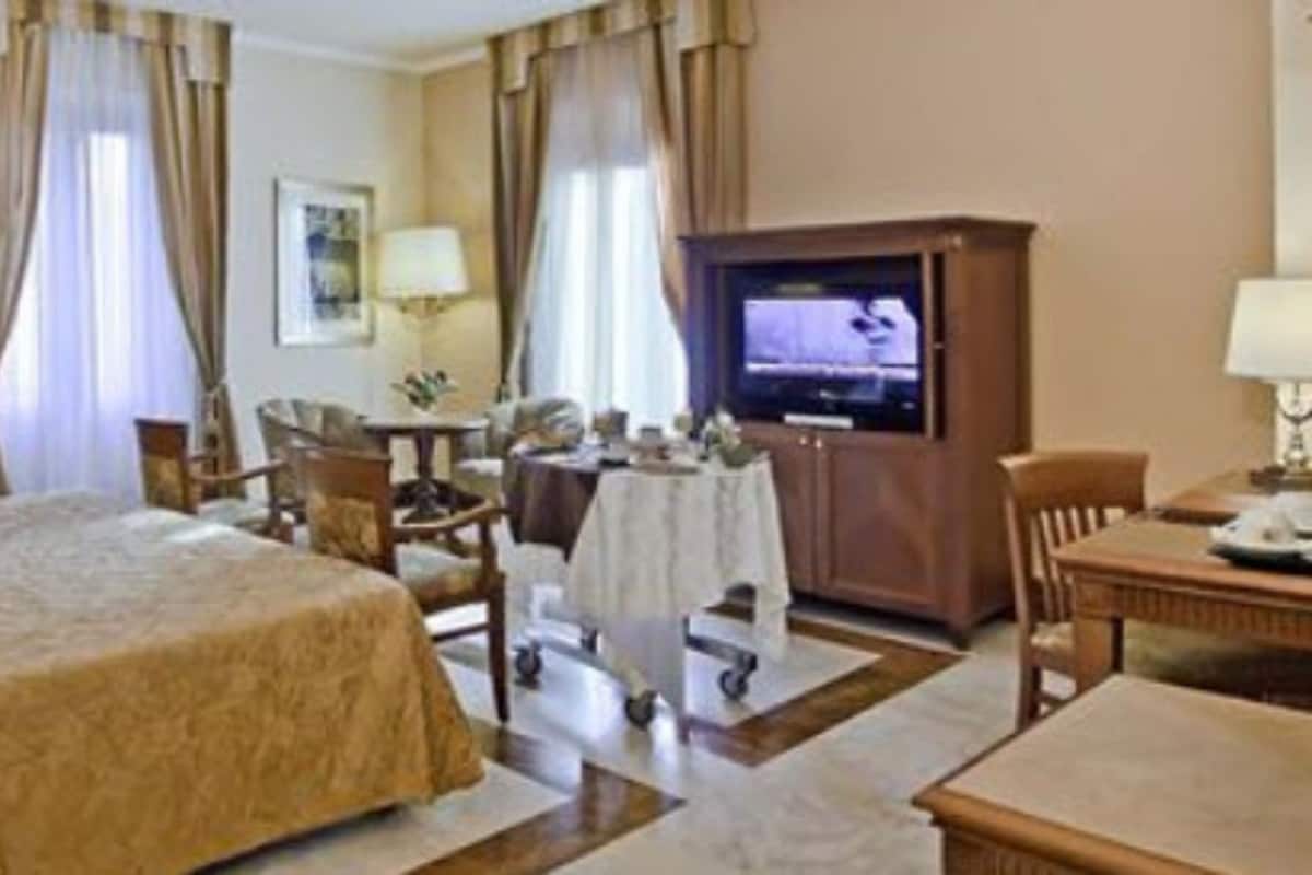 A hotel room with a large bed, a desk with a television, a chair, and a table with a lamp.