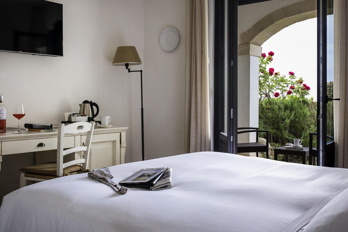 Habitación de hotel con cama, escritorio, silla, lámpara y ventana con vista a flores.