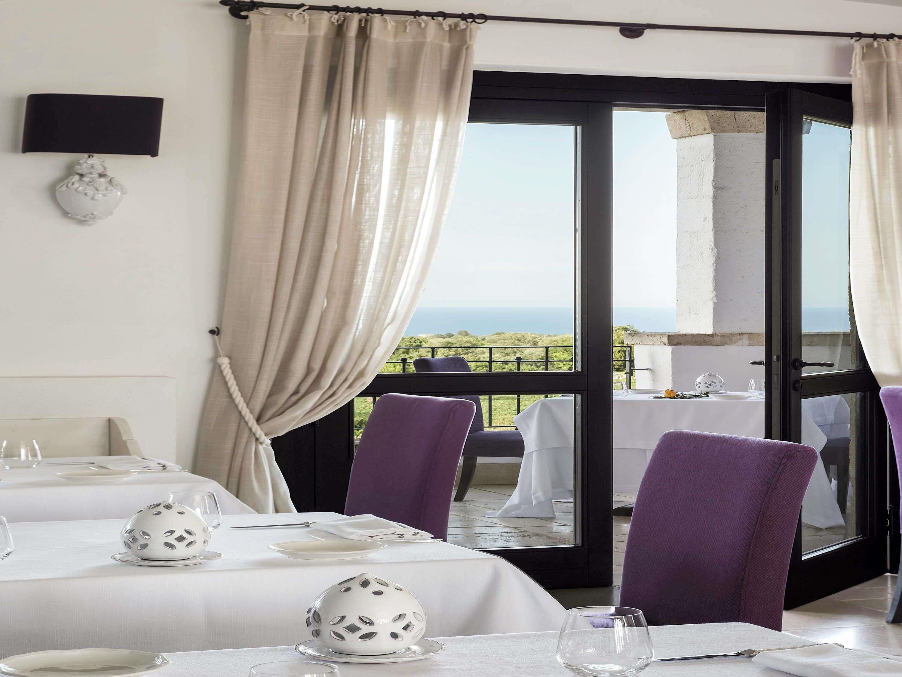 A dining area with tables set for two, a view of the sea, and purple chairs.