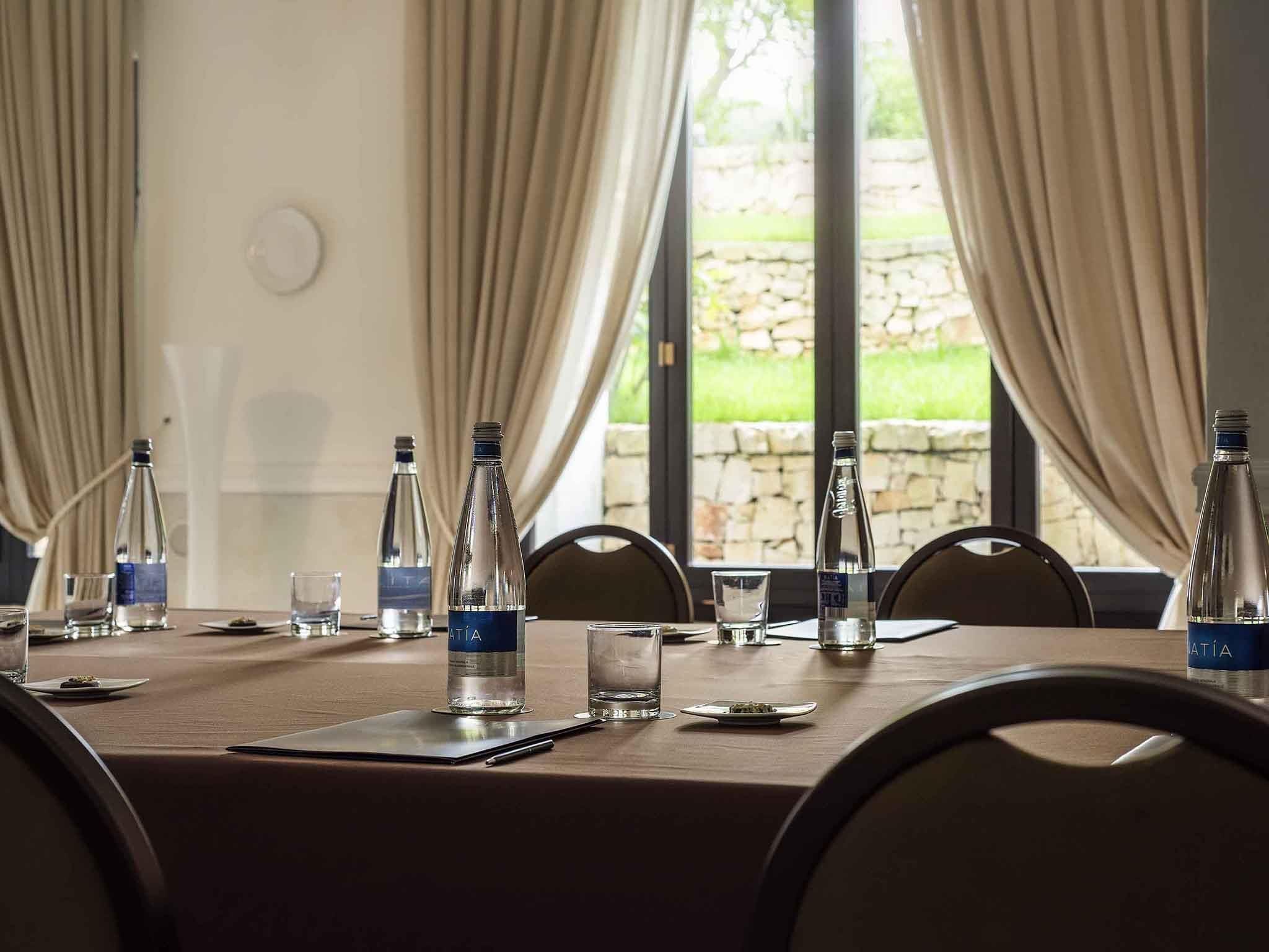 Una mesa de conferencias con botellas de agua, vasos y platos, frente a un fondo de cortinas y una pared de piedra.