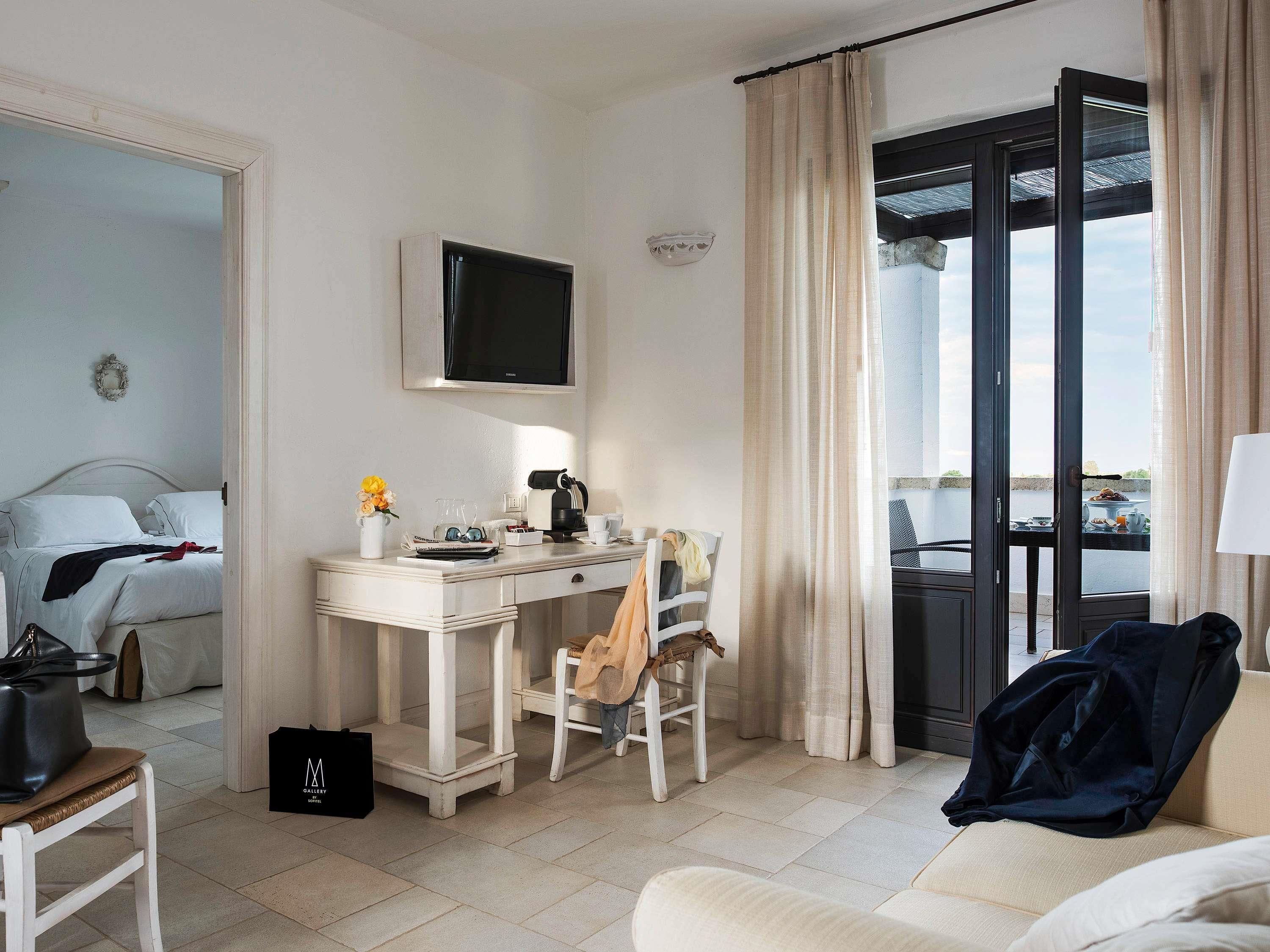Una camera d'albergo con un letto, una scrivania con un computer, una sedia, una finestra con tende e vista sull'esterno.
