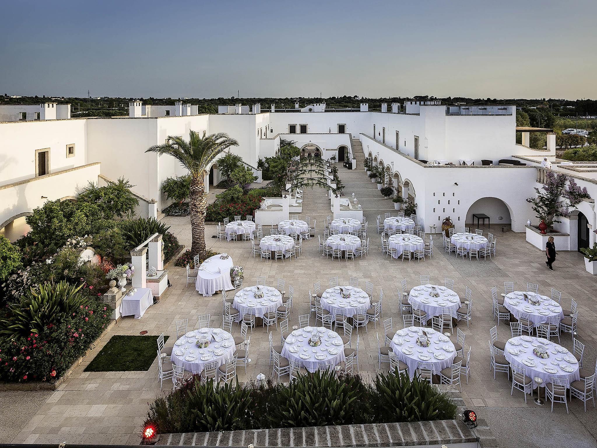 An outdoor dining area with round tables and chairs, surrounded by greenery and white walls.