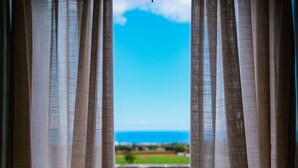 Vista a través de cortinas translúcidas a un balcón con vistas a un paisaje verde y un cielo azul despejado.