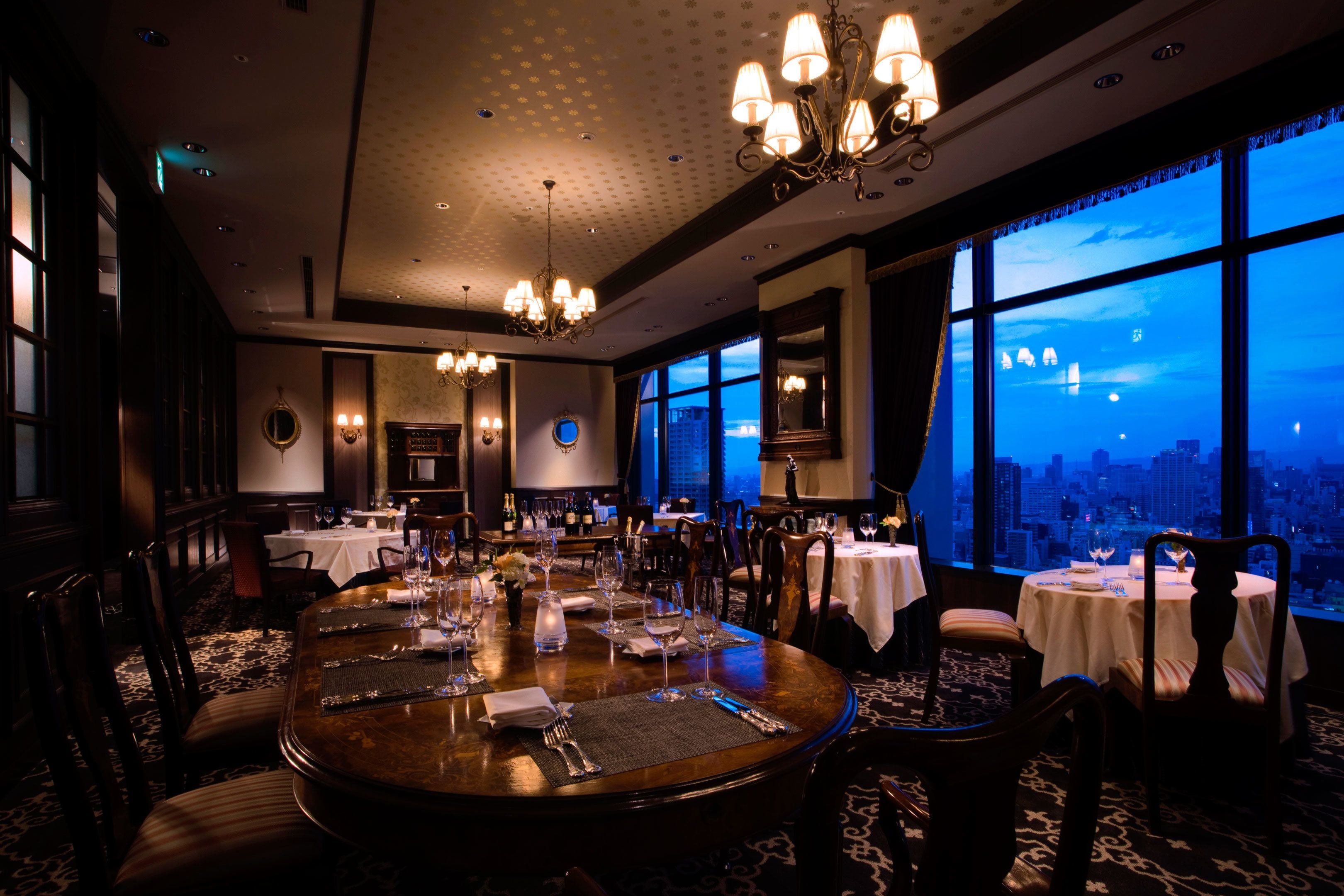 A dimly lit restaurant with round tables set for dining, large windows offering a city view, and classic chandeliers.