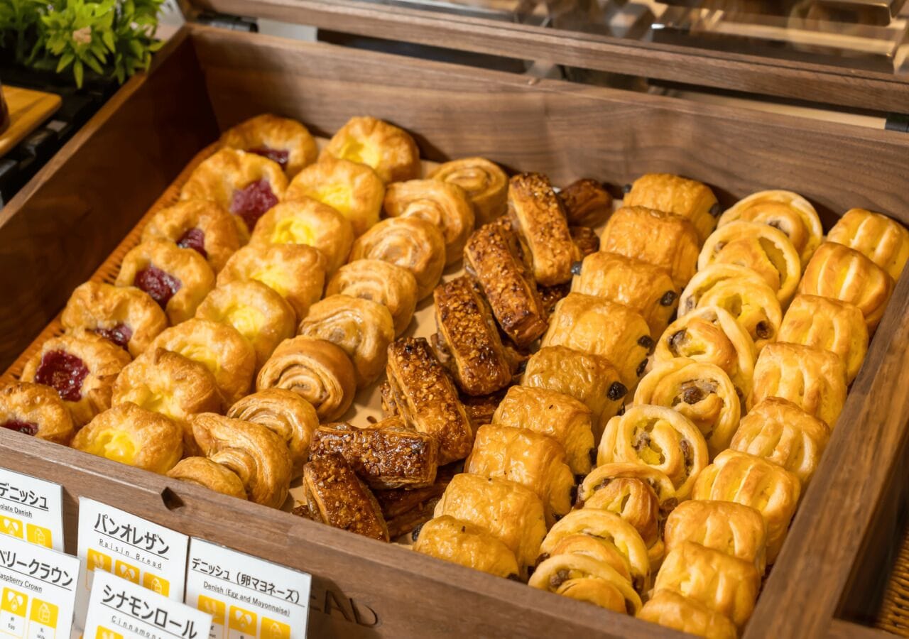 A wooden display case filled with various pastries, including croissants and Danish pastries, with labels in a foreign language.