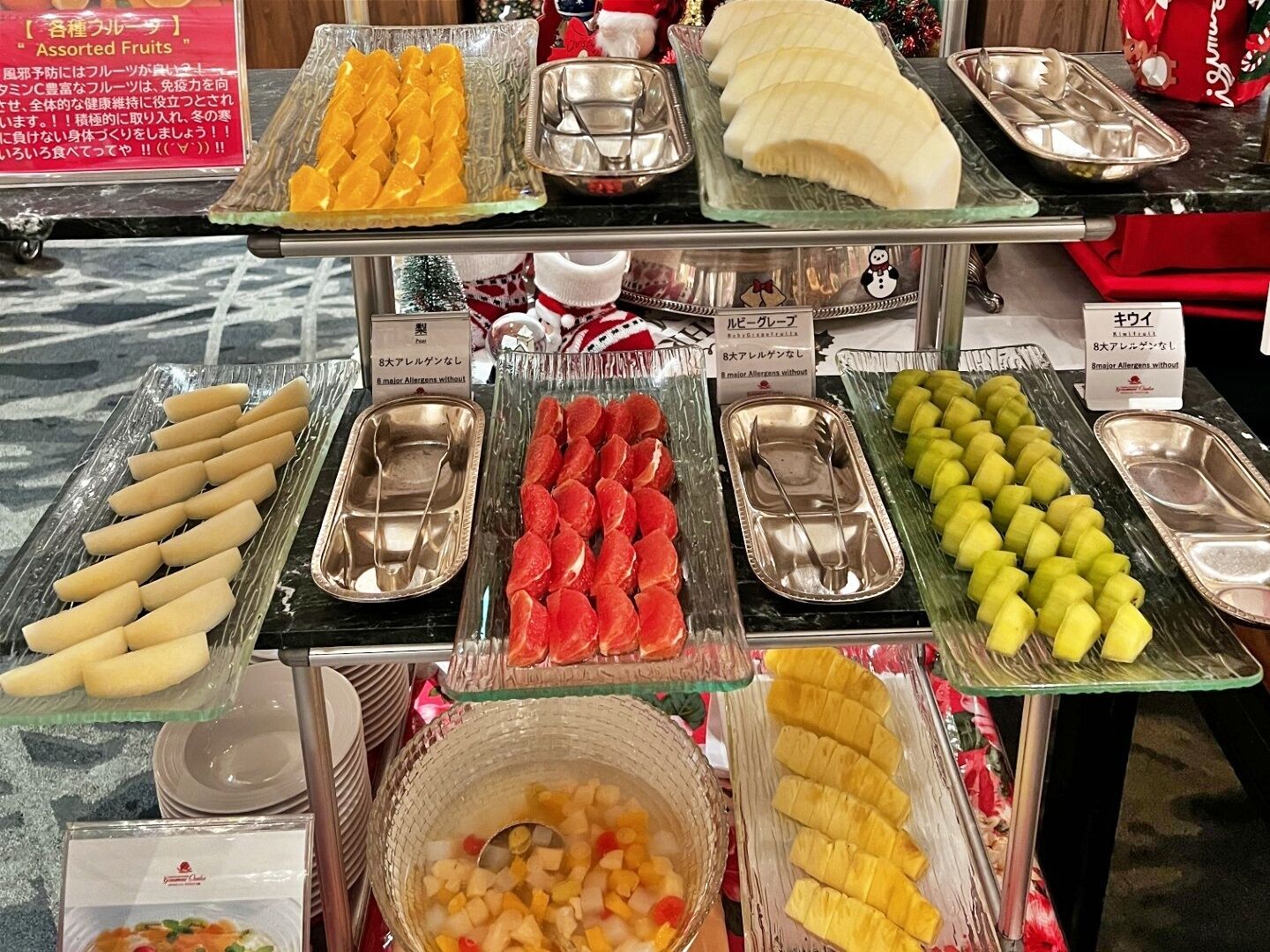 Un bufé con diversas frutas y verduras, incluyendo plátanos, naranjas y pepinos, dispuestas en bandejas con utensilios para servir.