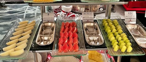 Un bufé con diversas frutas y verduras, incluyendo plátanos, naranjas y pepinos, dispuestas en bandejas con utensilios para servir.