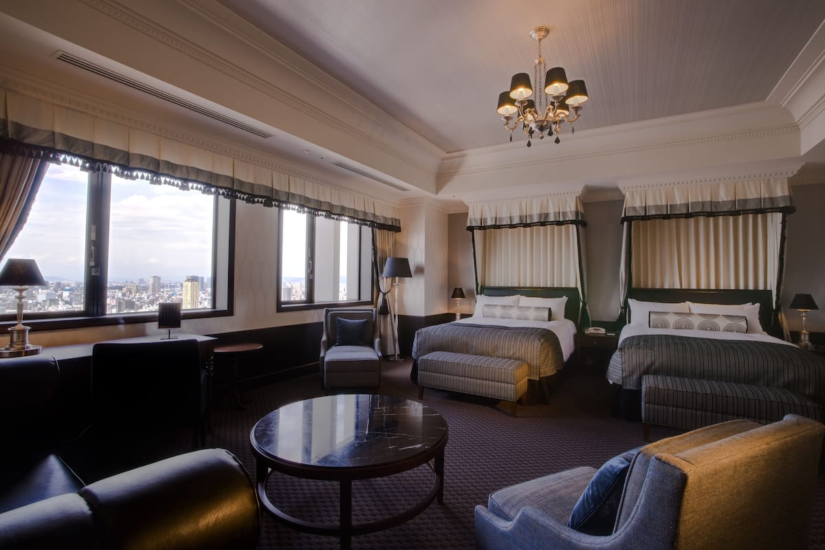 A hotel room with a large bed, a smaller bed, a sofa, a round table, and a view of the city.