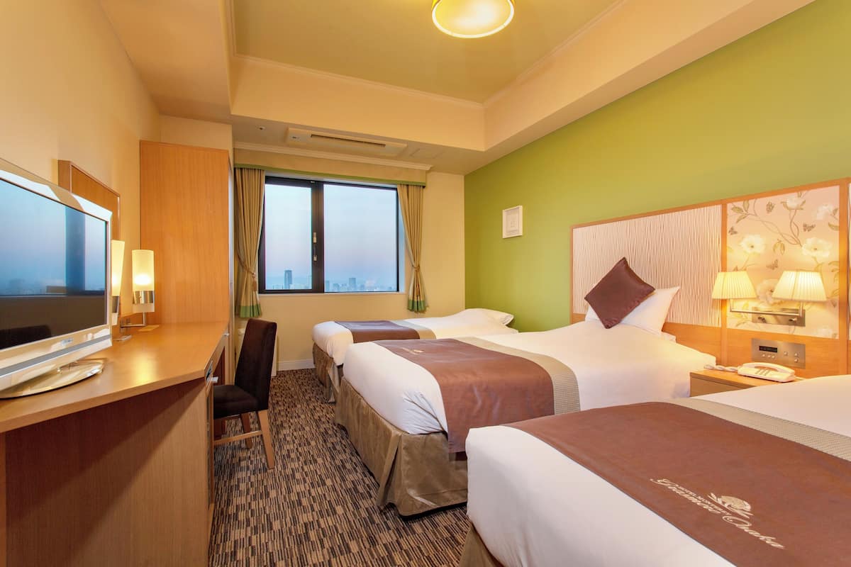 A hotel room with three beds, a desk with a TV, a window with a view, and a green accent wall.