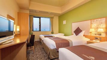 A hotel room with three beds, a desk with a TV, a window with a view, and a green accent wall.