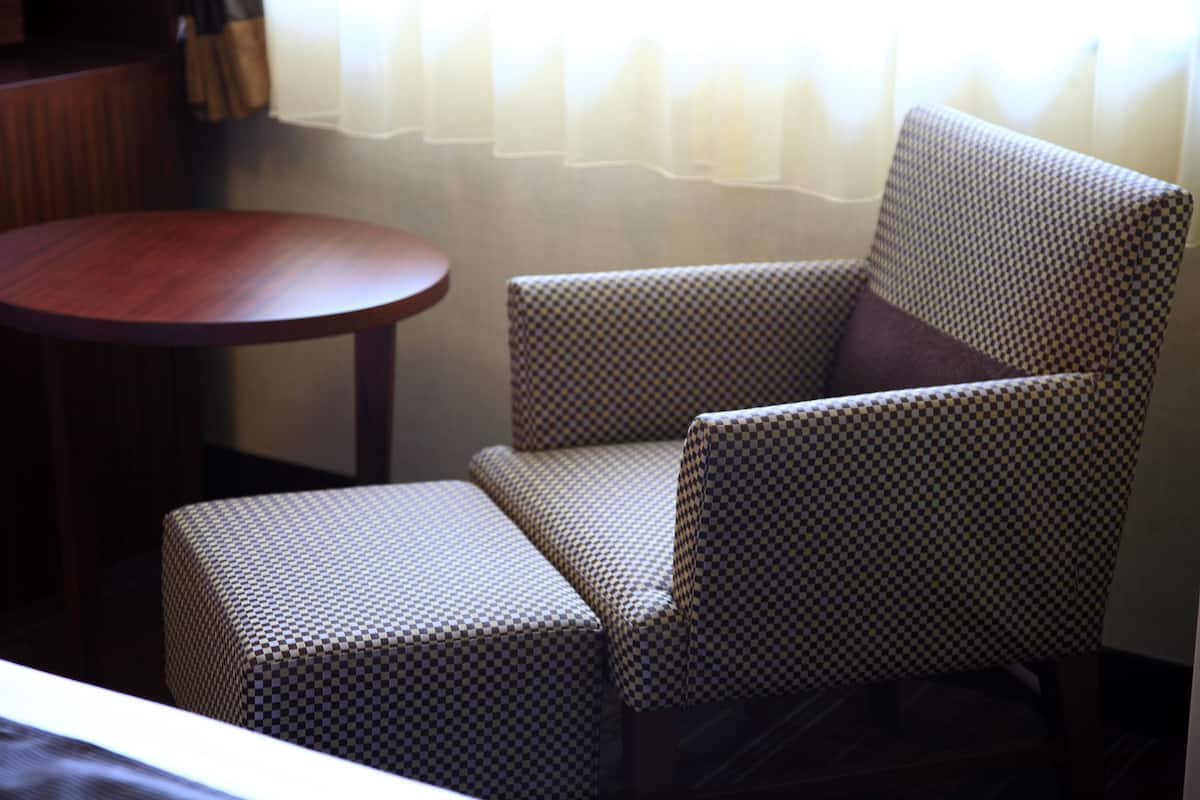A checkered armchair with a matching ottoman, a round wooden table, and a window with sheer curtains.