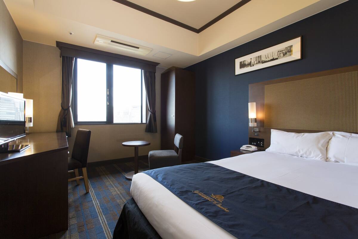 A hotel room with a large bed, a desk, a chair, a small table, and a window with curtains.