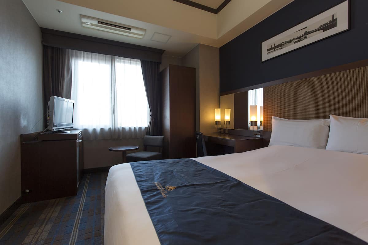 A hotel room with a large bed, a desk with a lamp, a chair, a small table, and a window with curtains.