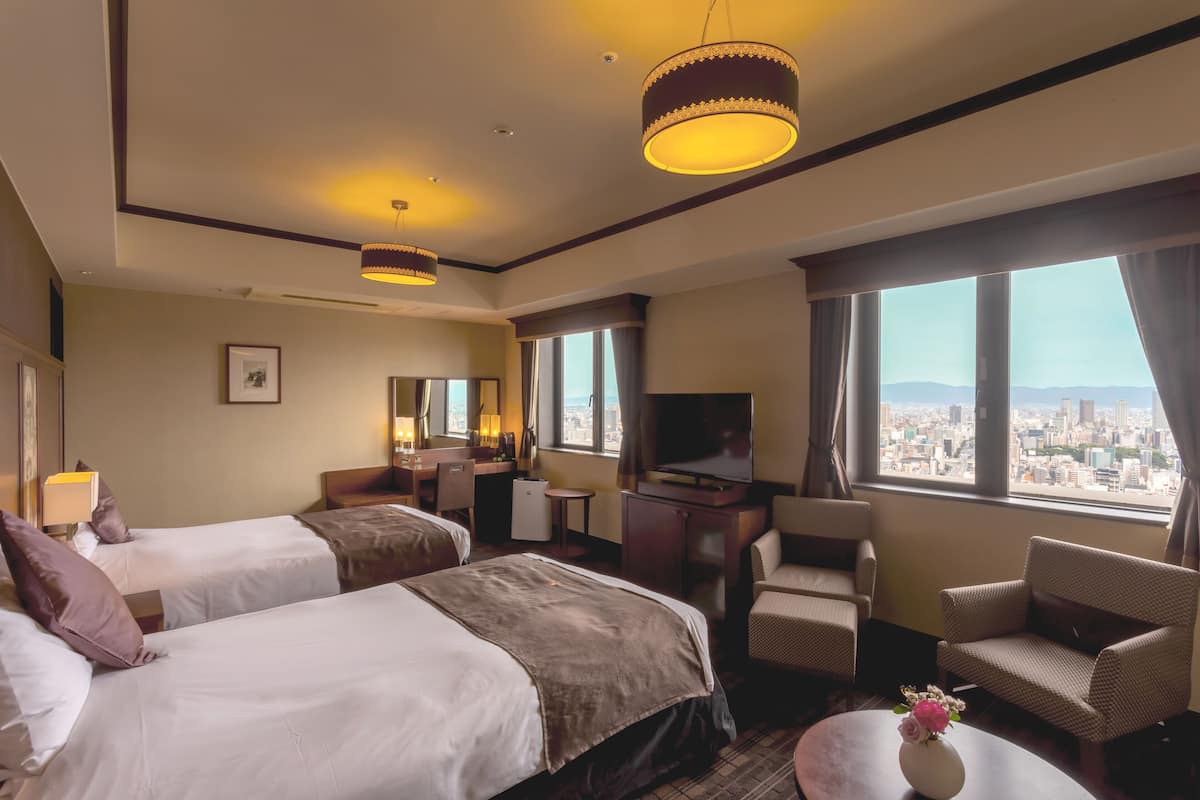 A hotel room with two beds, a desk, a chair, a TV, and a large window with a city view.