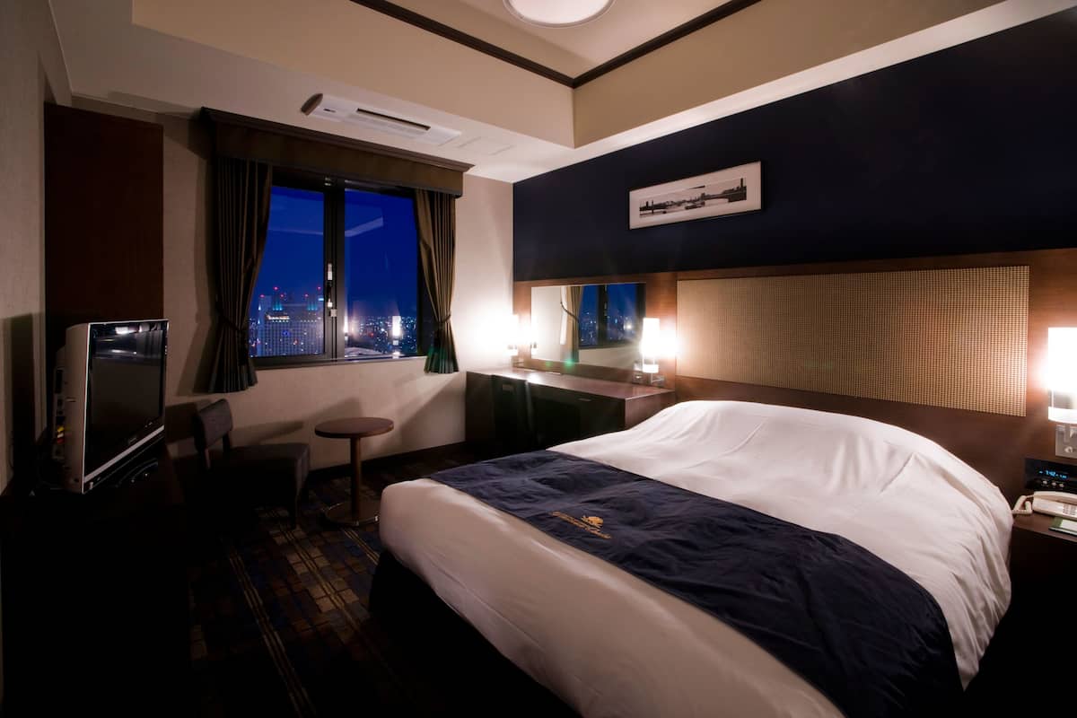 A hotel room with a large bed, a desk with a chair, a TV, and a view of the city at night.