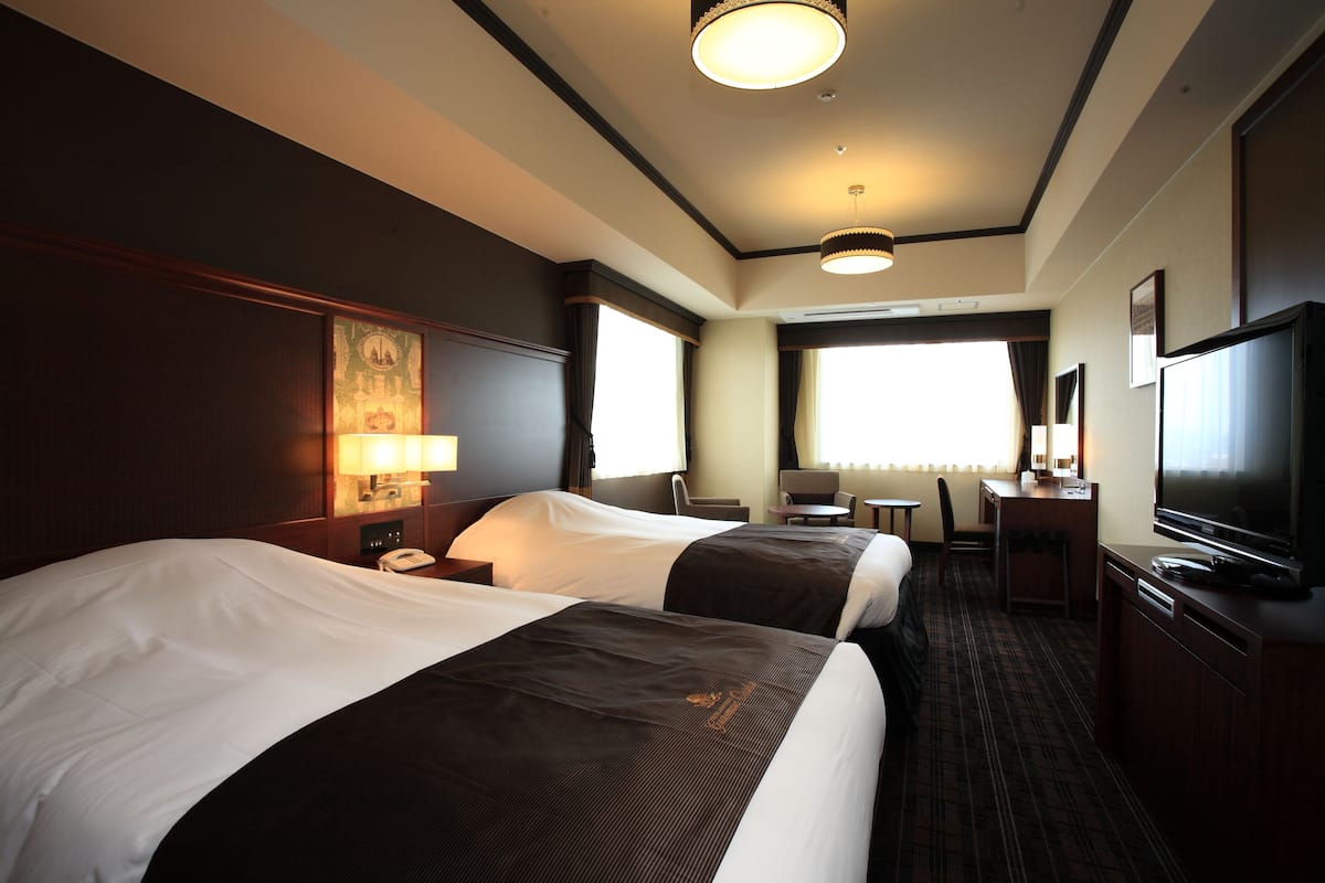 A hotel room with two beds, a desk, a chair, a television, and a large window.