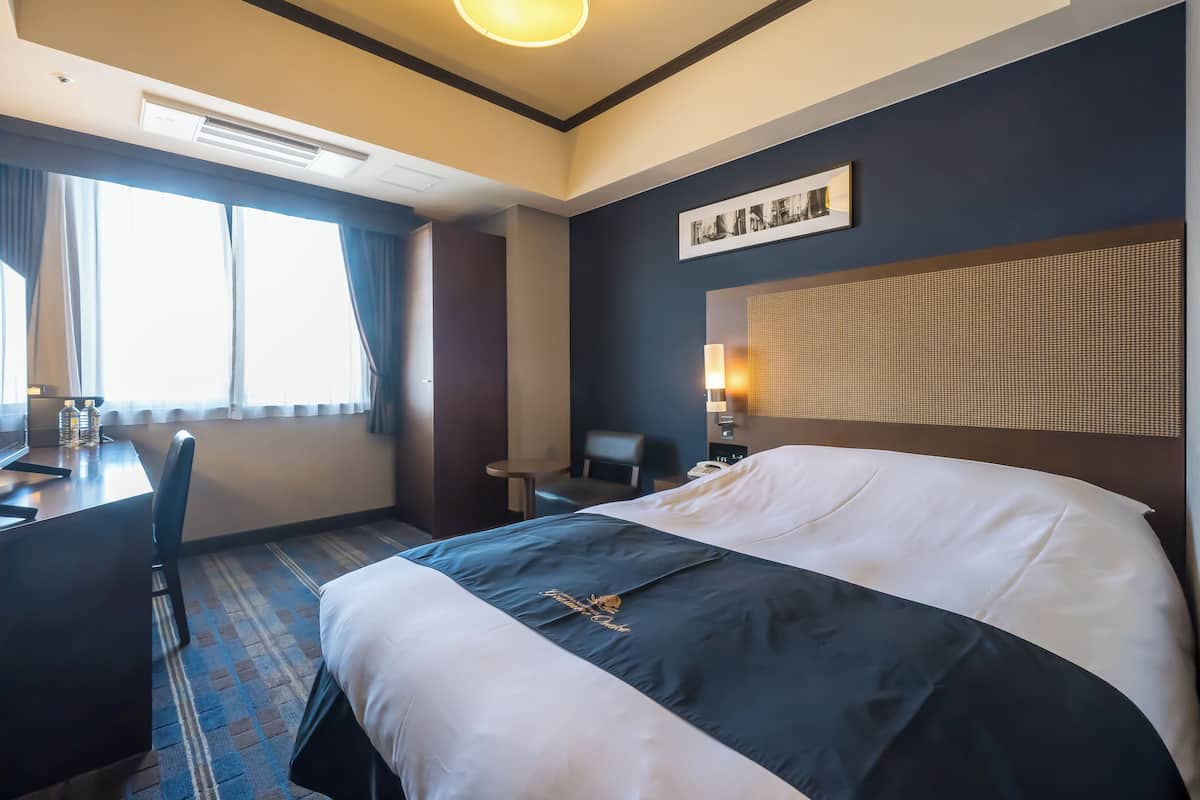 A hotel room with a large bed, a desk, and a chair. There is a window with curtains, a wall-mounted lamp, and a framed picture above the bed.