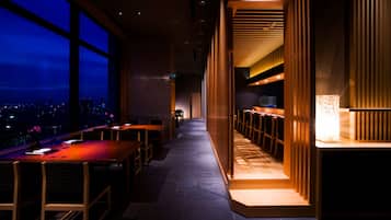 A modern restaurant with a long counter, wooden chairs, and a view of the city at night.