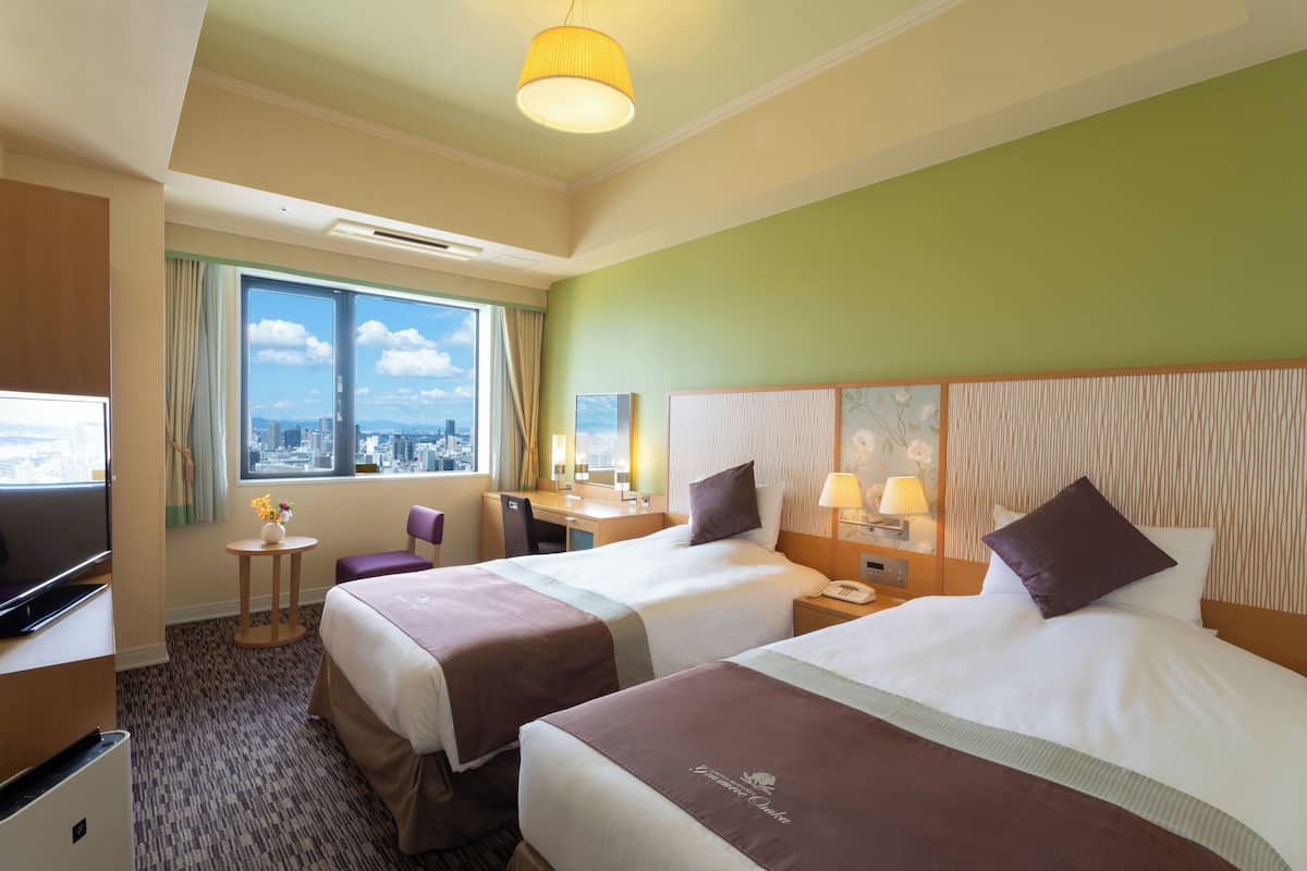 A hotel room with two beds, a desk, a chair, a TV, and a window with a city view.
