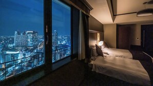 Hotellihuone, jossa on kaksi sänkyä, suuri ikkuna, josta on näkymä kaupunkiin, ja kattotuuletin.