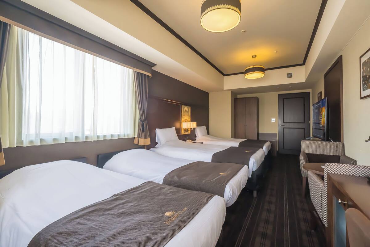 A hotel room with three beds, a desk, a chair, a television, and a door leading to a kitchenette.