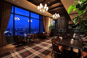 A dining area with a view of the city at dusk, featuring a plaid carpet, wooden furniture, and a chandelier.