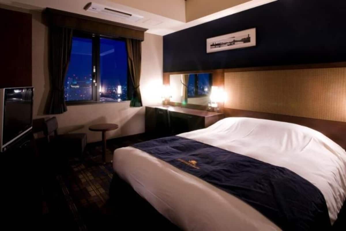 A hotel room with a bed, a desk, a chair, a TV, and a window with a city view.